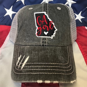 May include: A black and white distressed trucker hat with a white mesh back. The hat features an embroidered red and white outline of the state of Georgia with the text "GA girl" in red and a small white heart.