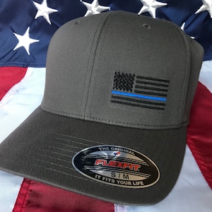 May include: A gray baseball cap with a black and blue American flag with a thin blue line embroidered on the front. The cap has a Flexfit label on the side with the text "The Original Flexfit S/M It Fits Your Life".