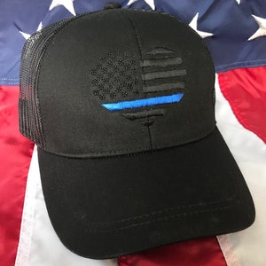 May include: Black trucker hat with an embroidered American flag design and a blue line. The flag is stylized in a circular shape. The hat has a mesh back and a curved peak.