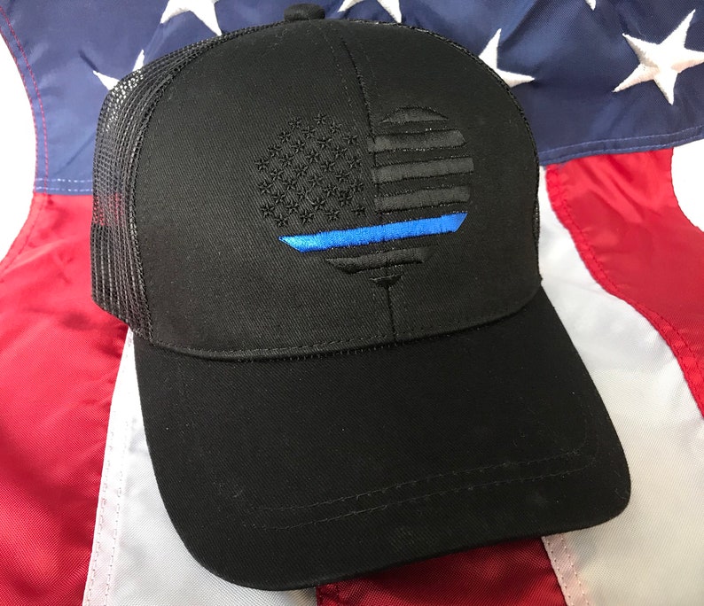 May include: Black trucker hat with an embroidered American flag design and a blue line. The flag is stylized in a circular shape. The hat has a mesh back and a curved peak.