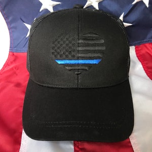 May include: Black baseball cap with a mesh back. The front has an embroidered design of the American flag with a blue line. The cap is set against a backdrop of the American flag.