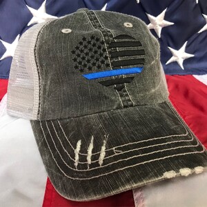 Puede incluir: Una gorra de camionero desgastada de color negro y gris con una malla blanca en la parte posterior. La parte delantera de la gorra presenta un coraz&oacute;n bordado con la bandera estadounidense con una l&iacute;nea azul en el centro.