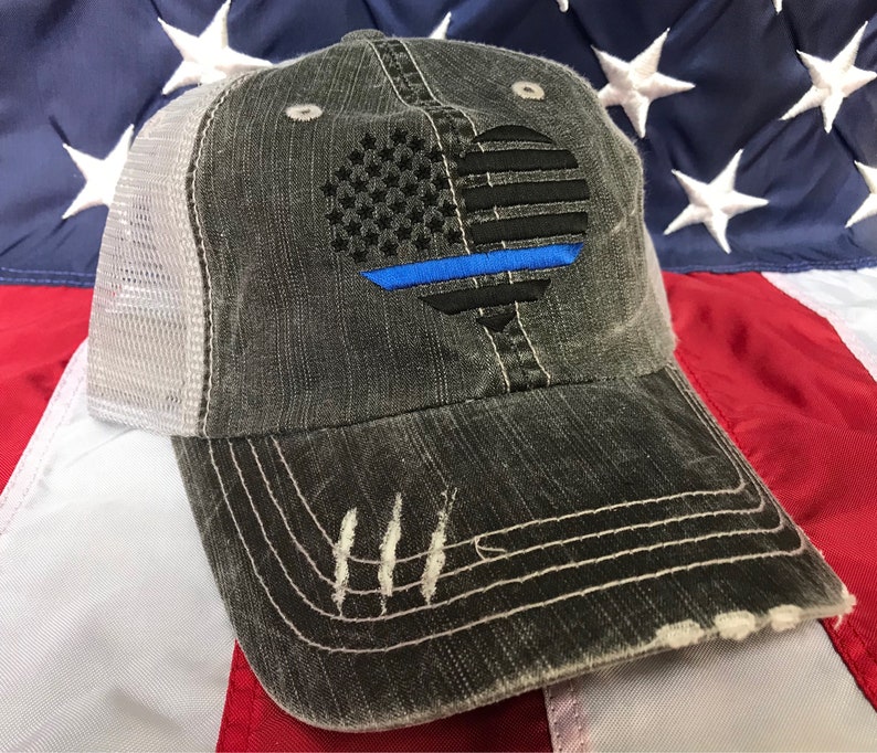Puede incluir: Una gorra de camionero desgastada de color negro y gris con una malla blanca en la parte posterior. La parte delantera de la gorra presenta un coraz&oacute;n bordado con la bandera estadounidense con una l&iacute;nea azul en el centro.