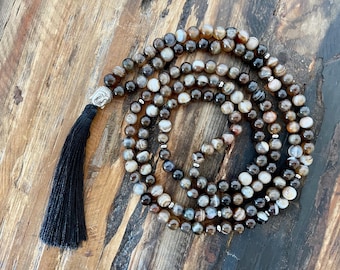 May include: A brown and grey agate mala with a silver Buddha head charm and a black tassel. The mala is made of 108 beads and is used for meditation and prayer.