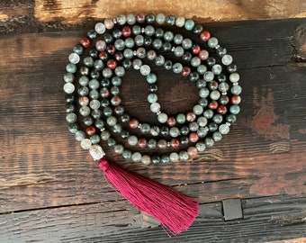 May include: A mala or prayer beads made of green, red, and white stones with a silver charm and a burgundy tassel.