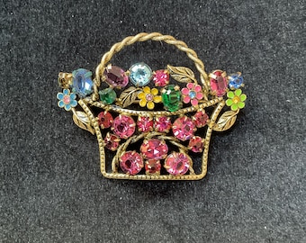 May include: A gold-tone brooch shaped like a basket overflowing with colorful rhinestones. The basket is decorated with green leaves and flowers.