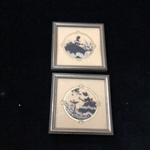 May include: Two framed silhouette art prints with a cream background and silver frames. The silhouettes are of women in dresses with gold accents.
