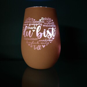 May include: A brown, egg-shaped glass candle holder with the words "du bist" in a heart shape, surrounded by positive adjectives in white script. The candle holder is illuminated from within, creating a soft glow.