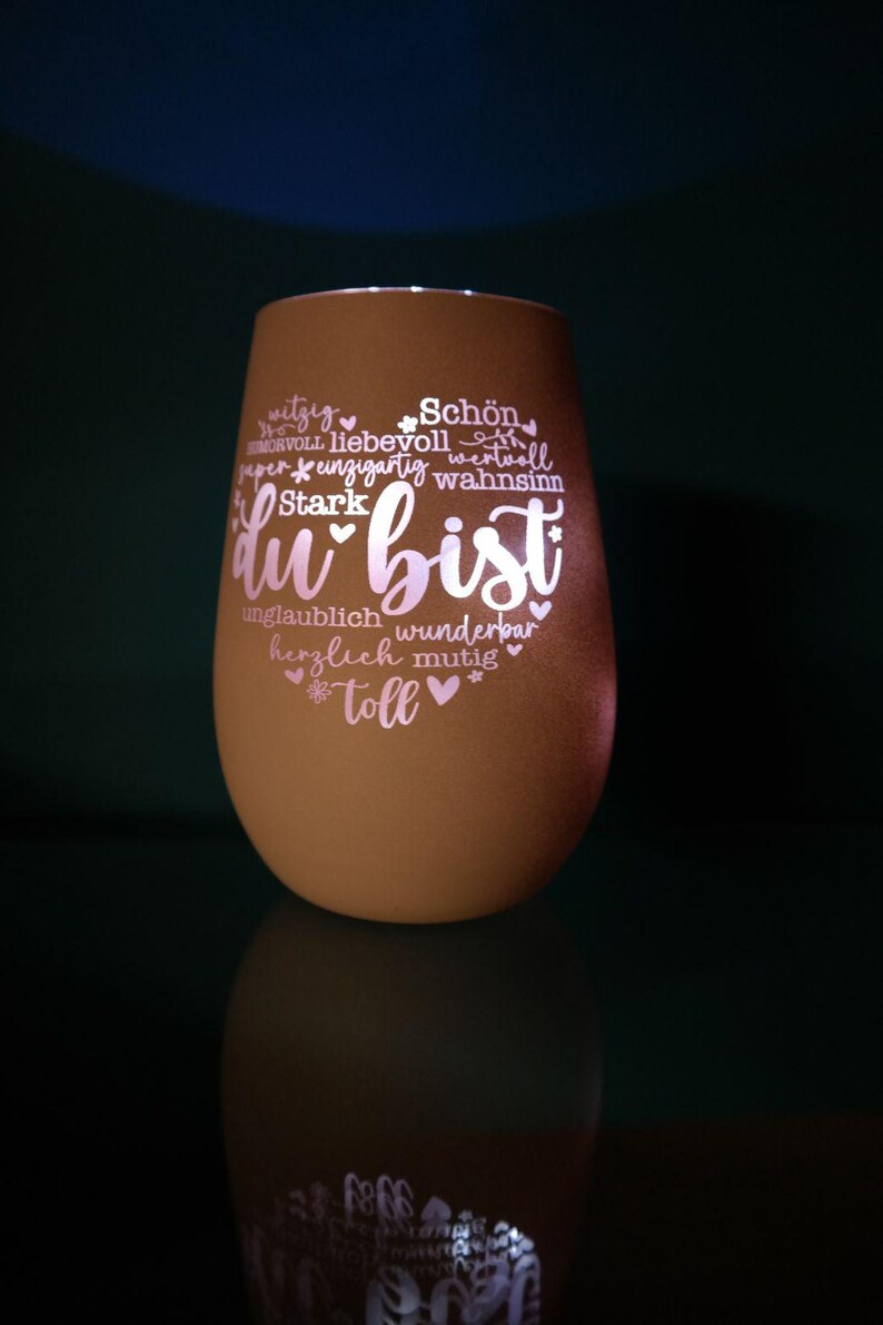May include: A brown, egg-shaped glass candle holder with the words "du bist" in a heart shape, surrounded by positive adjectives in white script. The candle holder is illuminated from within, creating a soft glow.