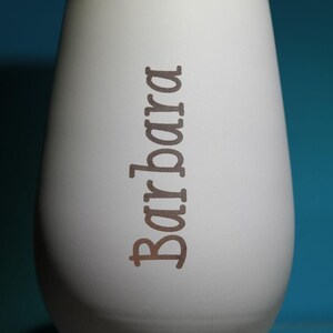 May include: A white, egg-shaped vase with the name "Barbara" printed vertically in a brown font. The vase has a smooth, matte finish and a slightly rounded base. The background is a solid teal color.