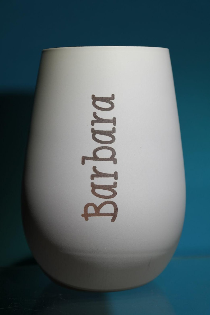 May include: A white, egg-shaped vase with the name "Barbara" printed vertically in a brown font. The vase has a smooth, matte finish and a slightly rounded base. The background is a solid teal color.