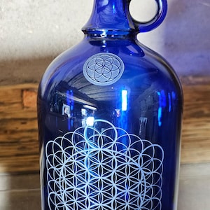 Könnte beinhalten: Eine kobaltblaue Glaskanne mit schwarzem Verschluss und Henkel. Die Kanne hat ein weißes geometrisches Muster auf der Vorderseite und eine kleinere Version auf der Schulter. Für Getränke oder Dekoration. Maße in Zentimetern.