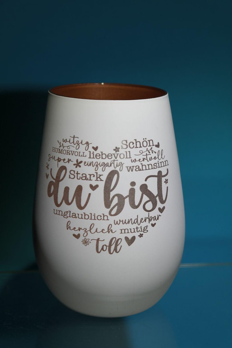 May include: White stemless wine glass with a brown rim. The glass features the German phrase "du bist" in a heart shape, surrounded by words like "beautiful" and "amazing".