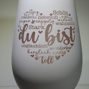 May include: A white stemless wine glass with the words "du bist" in a copper color, surrounded by positive adjectives in German. The glass has a smooth, matte finish and a rounded shape, perfect for a gift.