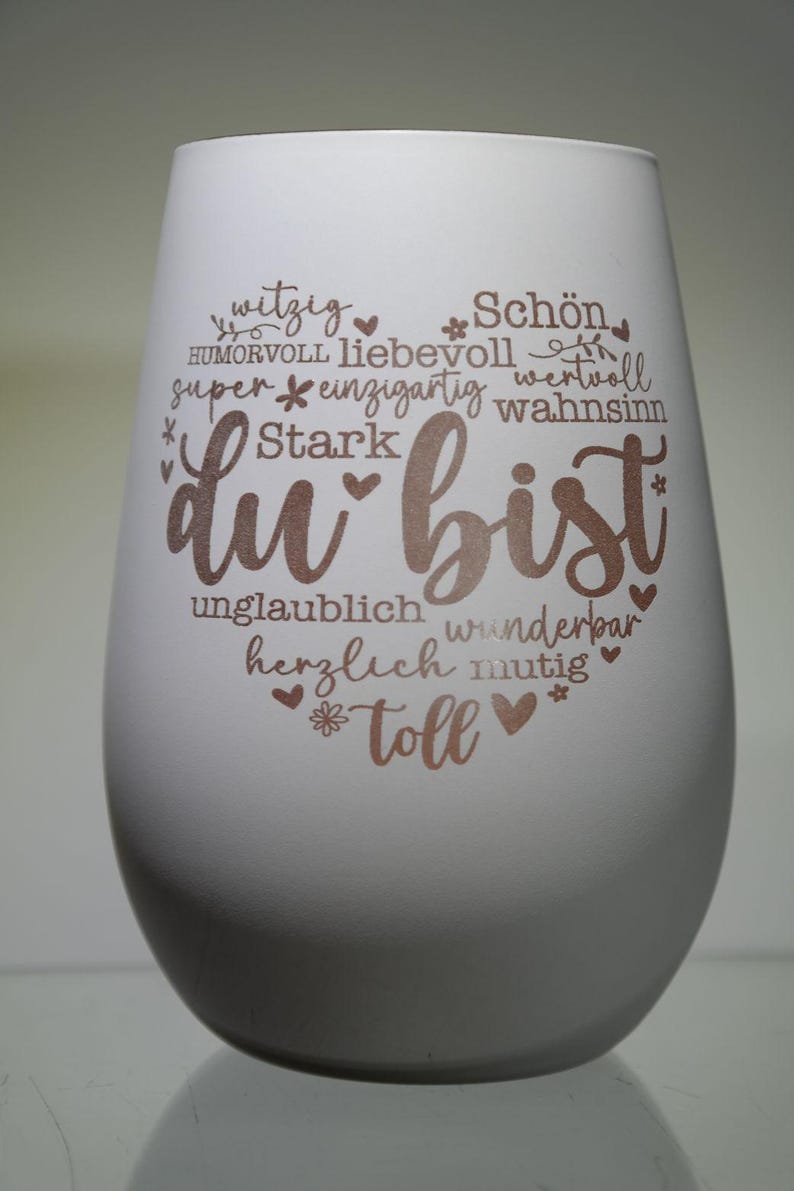 May include: A white stemless wine glass with the words "du bist" in a copper color, surrounded by positive adjectives in German. The glass has a smooth, matte finish and a rounded shape, perfect for a gift.