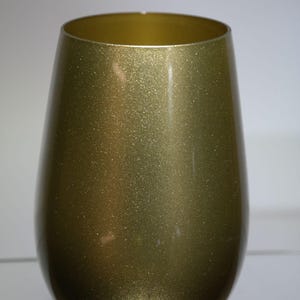 May include: A stemless wine glass with a metallic gold finish. The glass has a rounded body and a wide opening. The surface of the glass has a glittery, textured appearance, reflecting light. The glass is designed for wine.