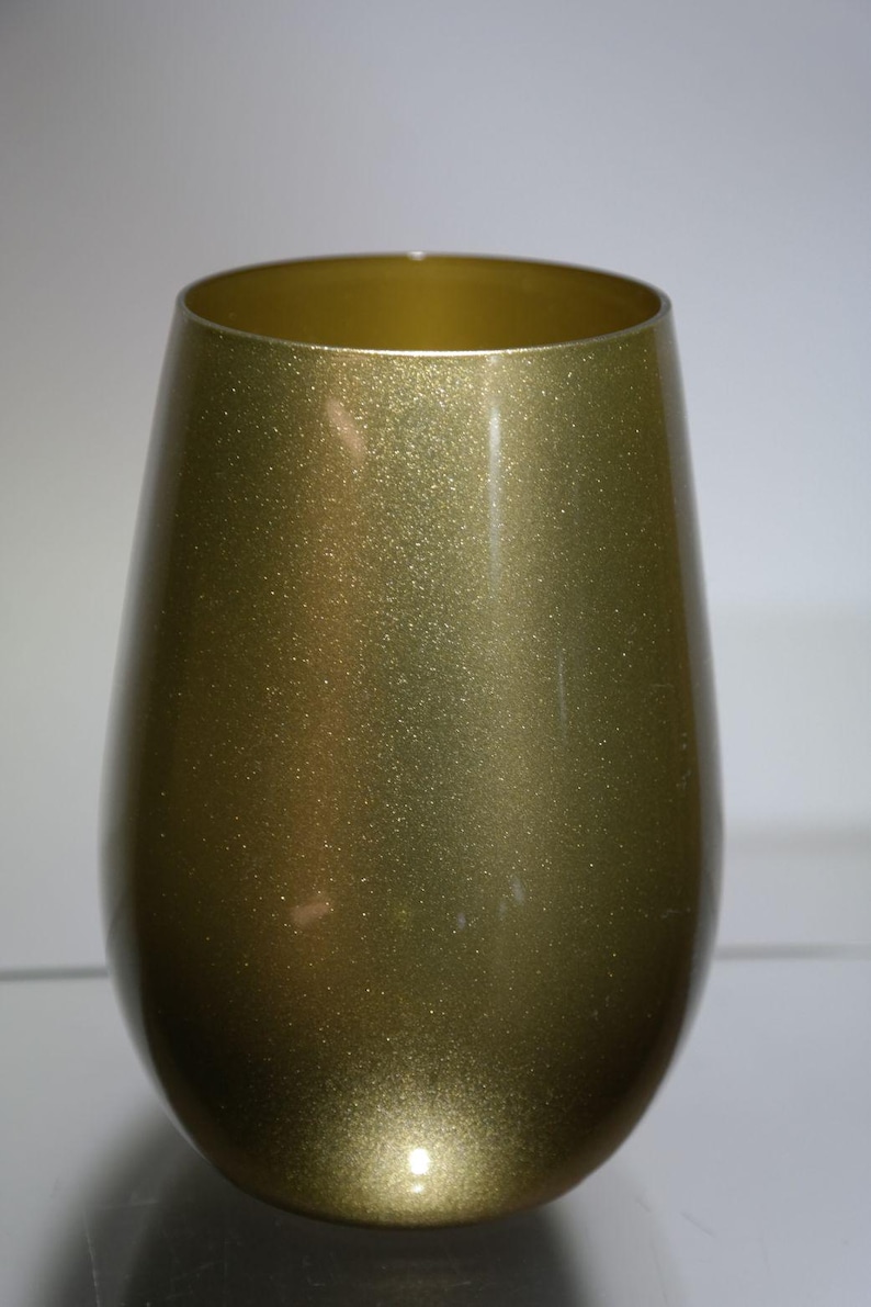 May include: A stemless wine glass with a metallic gold finish. The glass has a rounded body and a wide opening. The surface of the glass has a glittery, textured appearance, reflecting light. The glass is designed for wine.