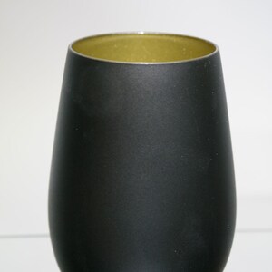 May include: A matte black stemless wine glass with a gold interior. The glass has a rounded base and a slightly flared rim. The interior has a metallic gold finish. The glass is designed for wine.