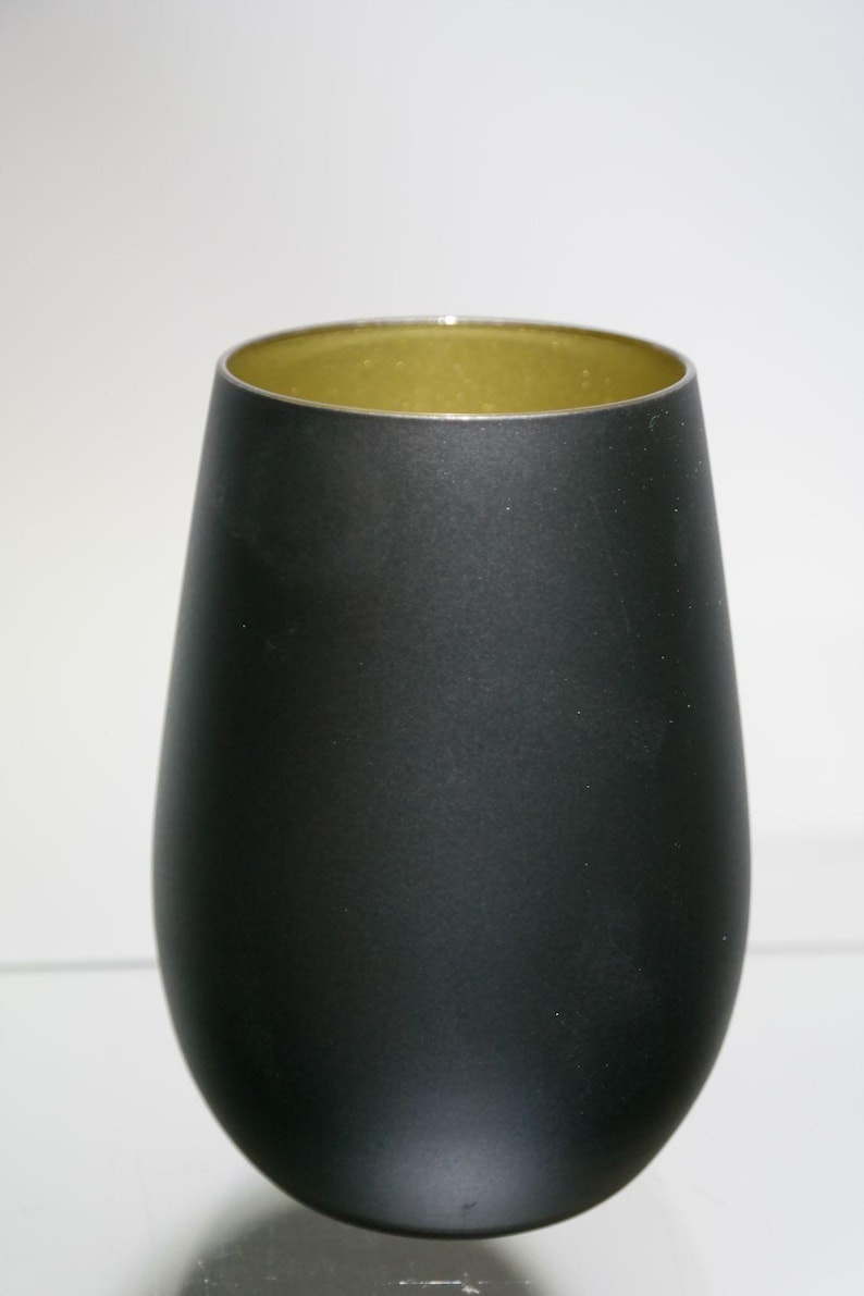 May include: A matte black stemless wine glass with a gold interior. The glass has a rounded base and a slightly flared rim. The interior has a metallic gold finish. The glass is designed for wine.
