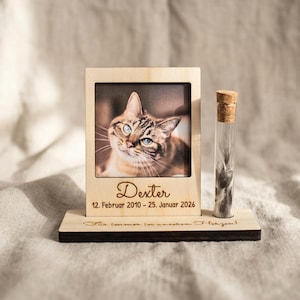 Könnte beinhalten: Ein Holzfotorahmen mit einem Foto einer Katze namens Dexter, mit den Daten 12. Februar 2010 - 25. Januar 2026. Daneben eine Glasampulle mit Korken. Aufschrift: "Für immer in unseren Herzen!"