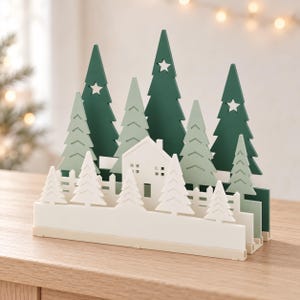 May include: A multi-layered wooden decorative display featuring a white house and fence in the foreground, surrounded by various shades of green pine trees. Some of the taller dark green trees have star-shaped cutouts near their tops.