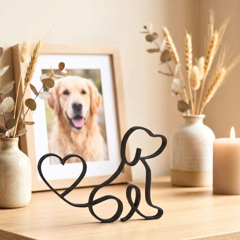 May include: A black metal dog silhouette with a heart-shaped tail sits on a wooden surface. A framed photo of a golden retriever is in the background, along with decorative vases and a lit candle.