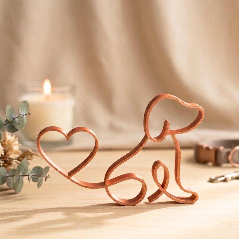 May include: A copper-colored metal sculpture of a dog with a heart-shaped tail. The dog is sitting, with its head and body formed by a continuous line. A lit candle and greenery are in the background. The sculpture is on a wooden surface.