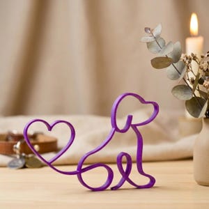 May include: A purple wire dog sculpture with a heart-shaped tail. The dog is sitting, with its head and body formed by a continuous wire. The sculpture is on a wooden surface, with a candle and a vase of flowers in the background.