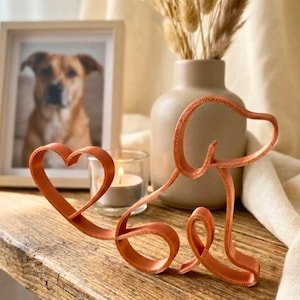 May include: A brown leather dog silhouette with a heart-shaped head and the word "Love" formed by the body. A framed photo of a dog, a lit candle, and a vase with dried flowers are in the background.
