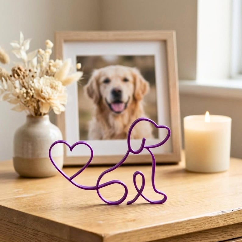May include: A purple wire dog sculpture with a heart-shaped tail sits on a wooden surface. A framed photo of a golden retriever is in the background, along with a lit candle and a vase of dried flowers. The sculpture is a decorative item.