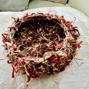 Peut inclure: Un panier rond en osier tress&eacute; de couleur marron, rouge et cr&egrave;me avec un bord effiloch&eacute;. Le panier est fait de laine et a une forme ronde.
