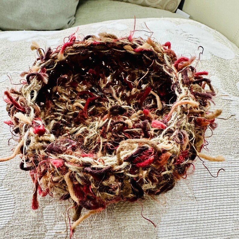 Peut inclure: Un panier rond en osier tress&eacute; de couleur marron, rouge et cr&egrave;me avec un bord effiloch&eacute;. Le panier est fait de laine et a une forme ronde.