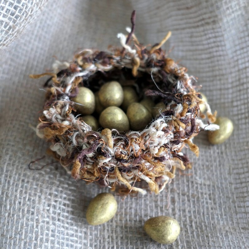 Peut inclure: Un nid en laine marron et blanc rempli d'&oelig;ufs en chocolat dor&eacute;s. Quatre autres &oelig;ufs sont &eacute;parpill&eacute;s sur une surface en toile de jute.