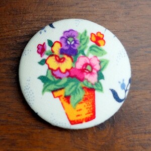 May include: A circular fabric button with a floral design. The image shows a colourful bouquet of pink, purple, yellow, and orange flowers in an orange pot against a white background. The button has a diameter of approximately 5 cm.