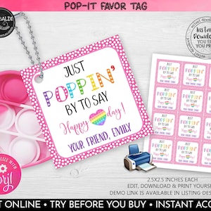 Puede incluir: Un juguete pop-it rosa con una etiqueta adjunta. La etiqueta es rosa con lunares blancos y dice "Just Poppin' By To Say Happy Day! Your Friend, Emily".
