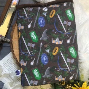 May include: A brown fabric book sleeve with a pattern of fantasy symbols, including swords, crowns, and bows. It has a button closure and a dark brown lining. The design incorporates green leaves and various fantasy elements.