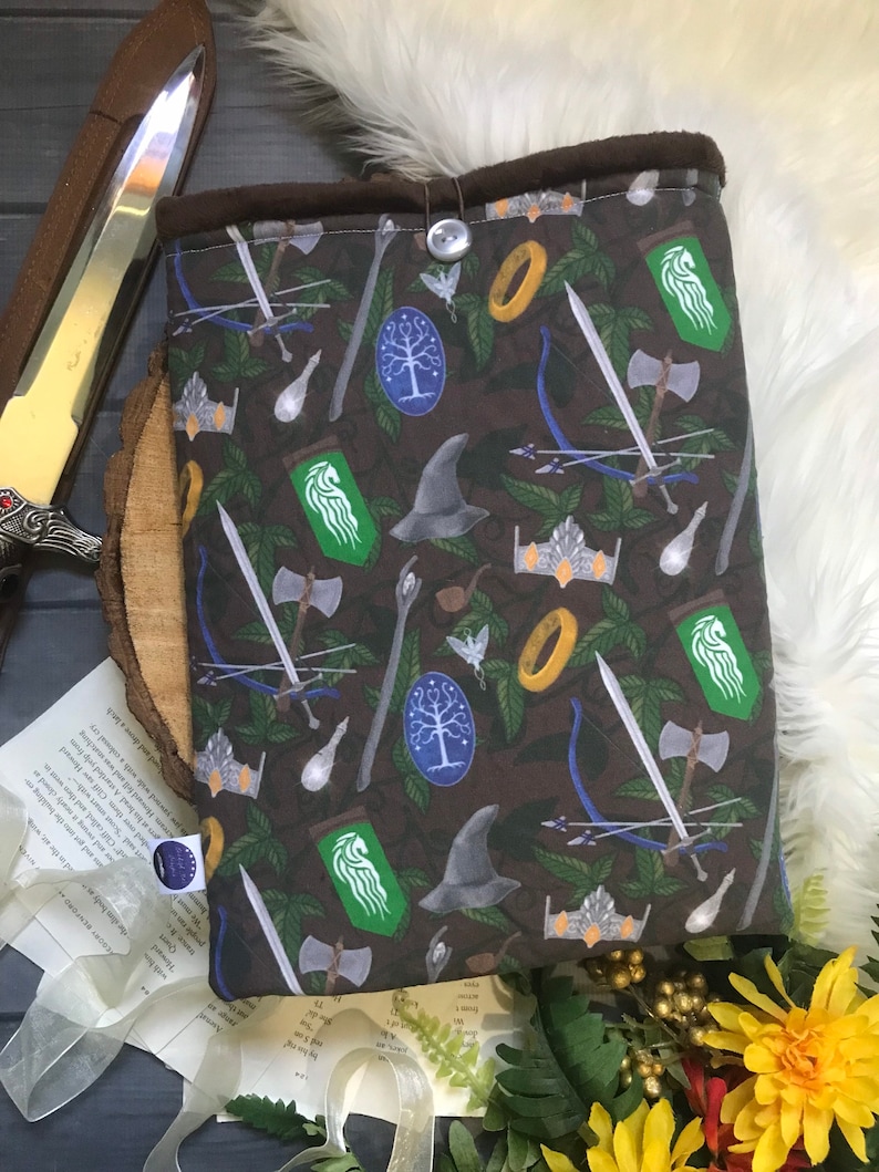 May include: A brown fabric book sleeve with a pattern of fantasy symbols, including swords, crowns, and bows. It has a button closure and a dark brown lining. The design incorporates green leaves and various fantasy elements.