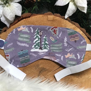 May include: A lavender sleep mask with a white elastic strap. The mask features a whimsical design with trees, signs, and text. The text includes phrases like "Courage, Dear Heart" and place names.