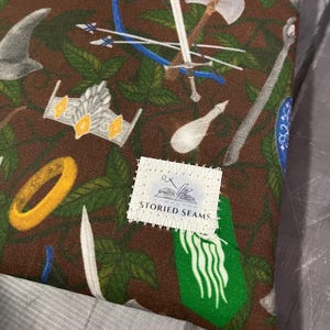 May include: A brown fabric featuring a repeating pattern of fantasy imagery, such as swords, bows, crowns, and rings. The design also includes green leaves and the text "STORIED SEAMS".