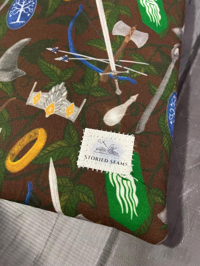 May include: A brown fabric featuring a repeating pattern of fantasy imagery, such as swords, bows, crowns, and rings. The design also includes green leaves and the text "STORIED SEAMS".