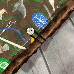 May include: Close-up of a brown fabric featuring a pattern of fantasy icons, such as axes, bows, and shields. The fabric is trimmed with a brown, fuzzy material and fastened with a button and elastic bands. The overall theme is fantasy.