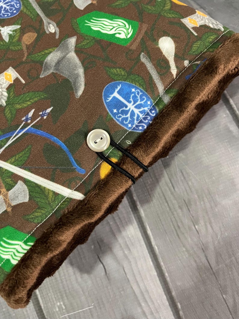 May include: Close-up of a brown fabric featuring a pattern of fantasy icons, such as axes, bows, and shields. The fabric is trimmed with a brown, fuzzy material and fastened with a button and elastic bands. The overall theme is fantasy.