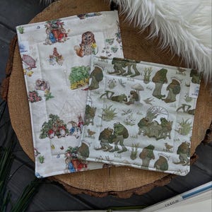 May include: Two white fabric squares with different designs. One square features a whimsical woodland scene with animals and plants. The other square features a repeating pattern of green frogs in a garden setting. Both squares have a white background and are stitched with a white thread.