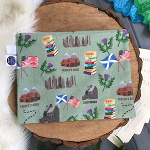 Może przedstawiać: Jasnozielona kosmetyczka zapinana na zamek z powtarzającym się wzorem ilustracji o tematyce szkockiej i amerykańskiej. Projekt zawiera amerykańską flagę, szkockie flagi, stosy książek oraz słowa "Fraser's Ridge" i "Lallybroch".