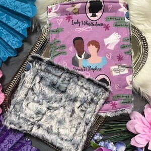 May include: A pink and white fabric square with a repeating pattern of hearts, flowers, and text that reads "Lady Whistledown", "Simon & Daphne", and "I am yours, always". The fabric is folded in half and has a gray and white fuzzy fabric square on top of it.