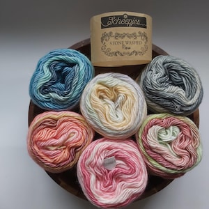 May include: Six round skeins of Scheepjes Stone Washed Flow yarn in various gradient colors including blue, yellow, pink, and grey. The yarn is arranged in a circular pattern with a tan paper label featuring the text Scheepjes Stone Washed Flow.