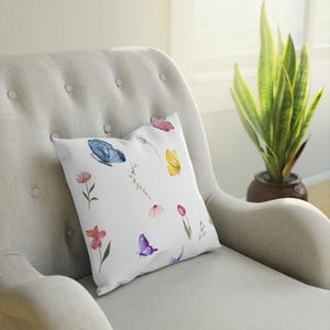 May include: A white throw pillow with a watercolor design of butterflies and flowers. The butterflies are blue, yellow, and purple. The flowers are pink, red, and yellow.