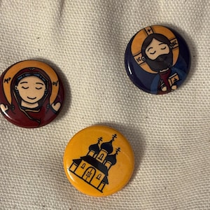May include: Three circular pinback buttons featuring cartoon-style illustrations. One button shows a figure in a red robe with a gold halo, another shows a figure in a blue robe with a gold halo, and the third is a yellow button with a black church.