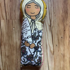 May include: A fabric doll with a serene face, wearing a white veil and a floral patterned dress. The doll has the text "Strong" and a quote printed on a paper. The doll is approximately 8 inches tall.