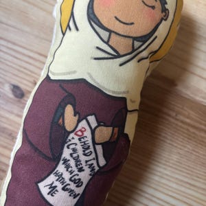 May include: A fabric doll with a cartoon illustration of a person in a maroon robe and white head covering. The doll holds a scroll with the text "BEHOLD I AND THE CHILDREN WHICH GOD HATH GIVEN ME". The doll has rosy cheeks and closed eyes, with a yellow halo.